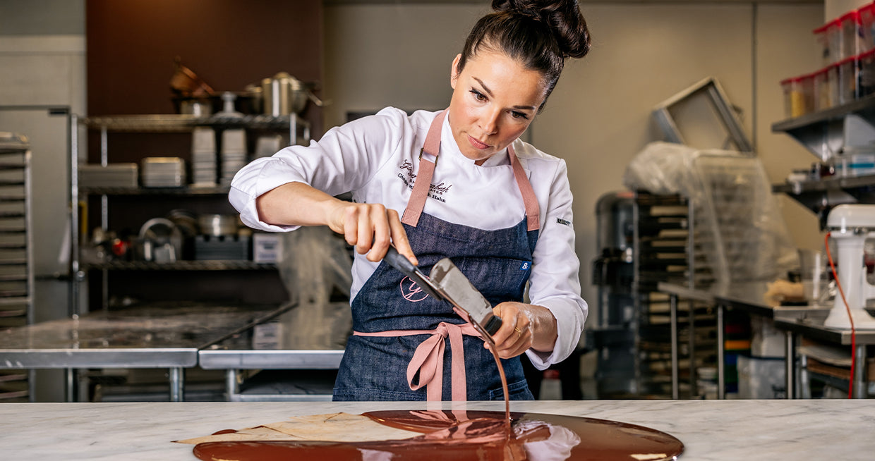 Gnger Elizabeth working with melted chocolate on mable ountertop and commercial kitchen in background. 