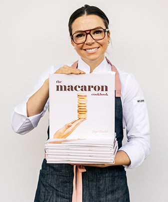 woman standing face forward holding stack of Macaron Cookbooks with top book upright showing cover in front of white background. 