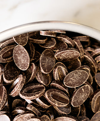 Close up of stainless steel bowl of bittersweet chocolate medallionswith marble counter in background. 
