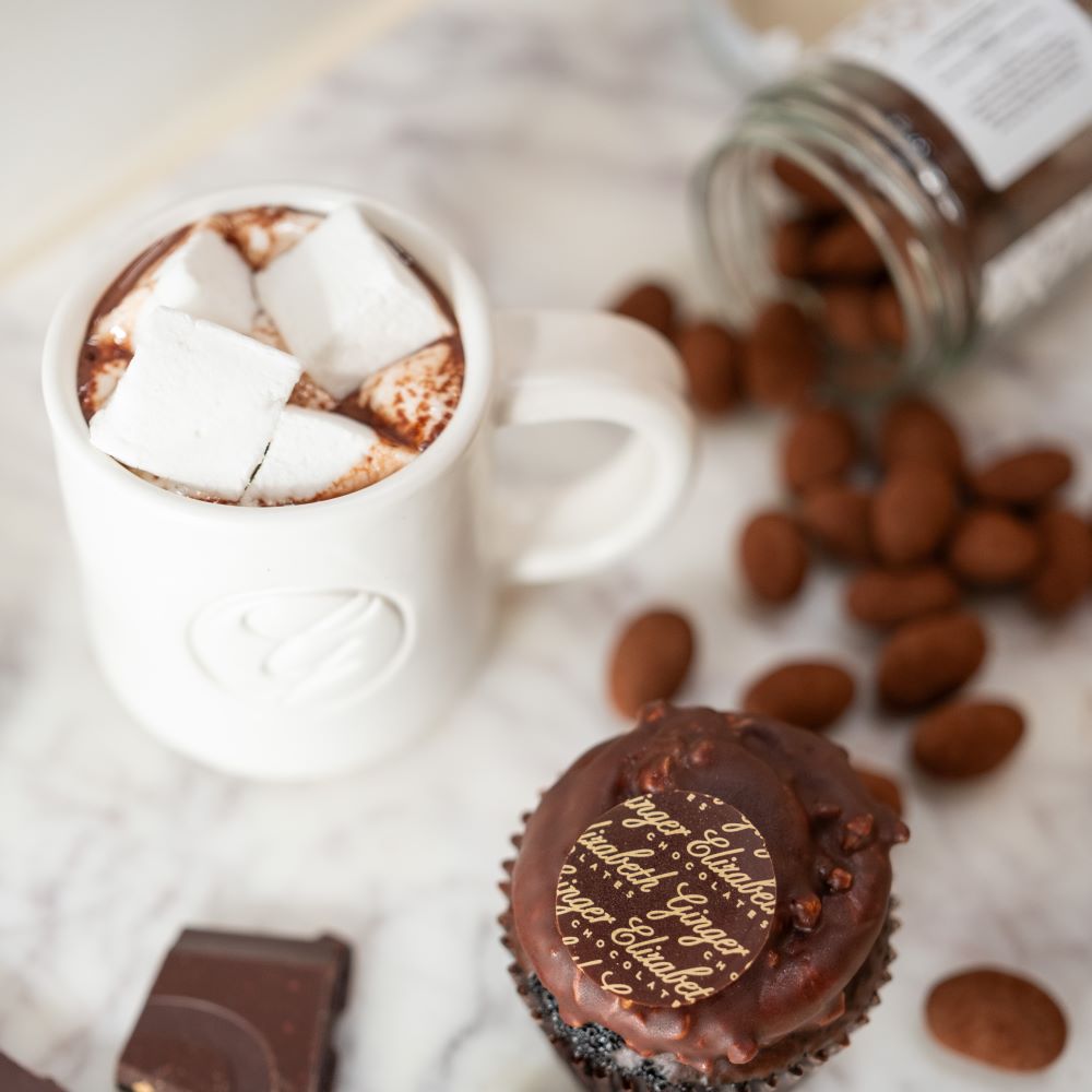 Mug of hot chocolate with marshmallows displayed with cupcake and open jar of candied chocolate almonds blurred in background