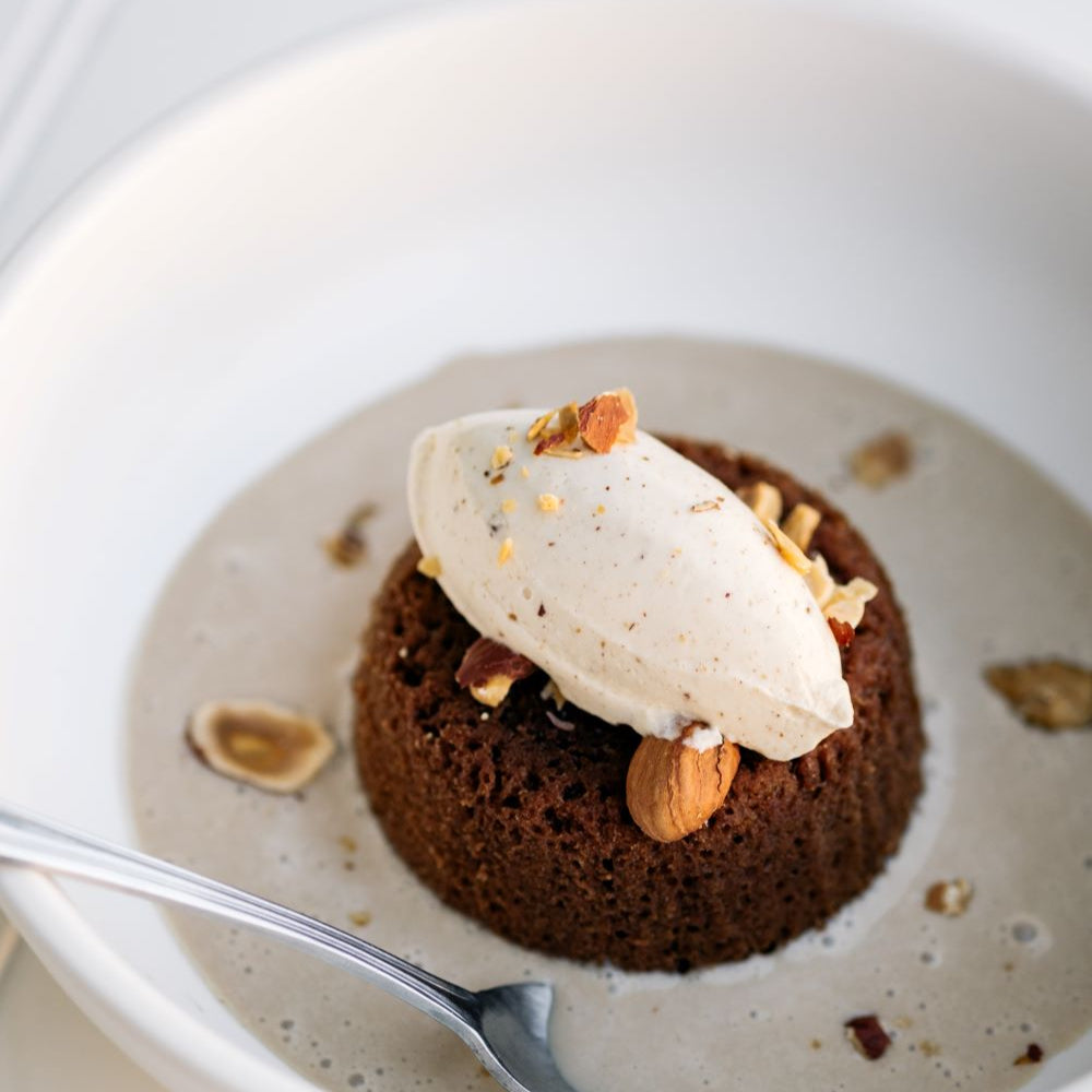 Ginger Elizabeth's Chocolate Molten Lava Cake. Individual servings in bowl with spoon,  cream anglaise, whipped cream and toasted hazelnuts on white bead board background .