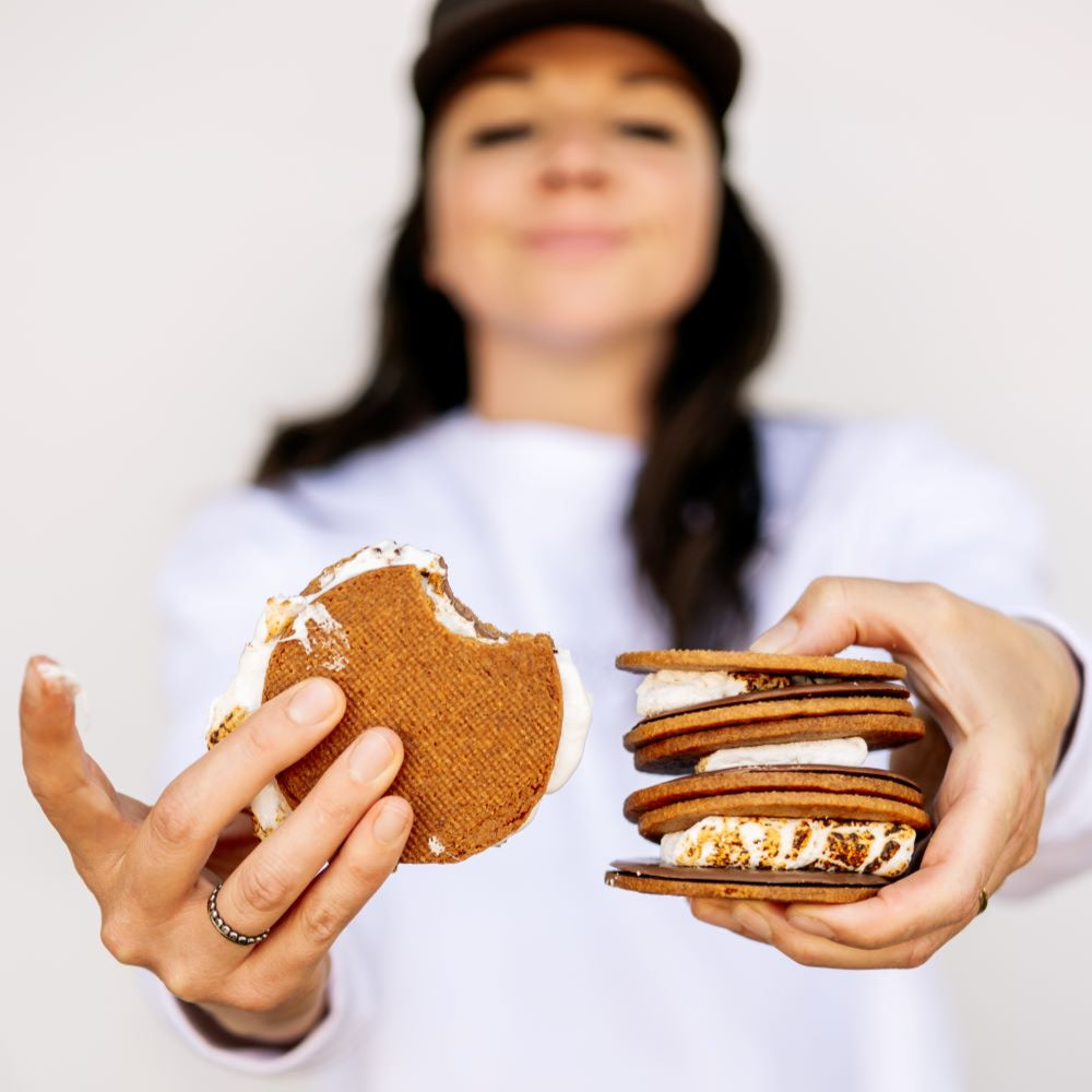Woman in brown logo hat, white top and jeans holding smores out in front of her blurred body.