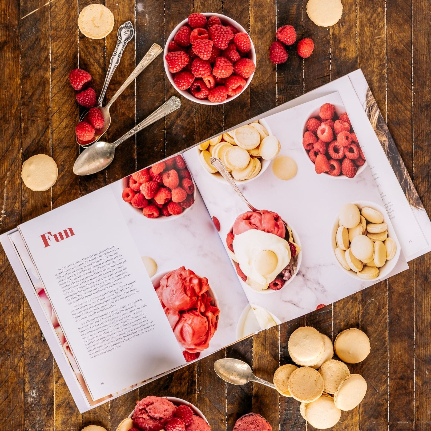 The Macaron Cookbook open to page showing raspberry ice cream, fresh raspberries and macaron cookies surround by same items on dark wood background