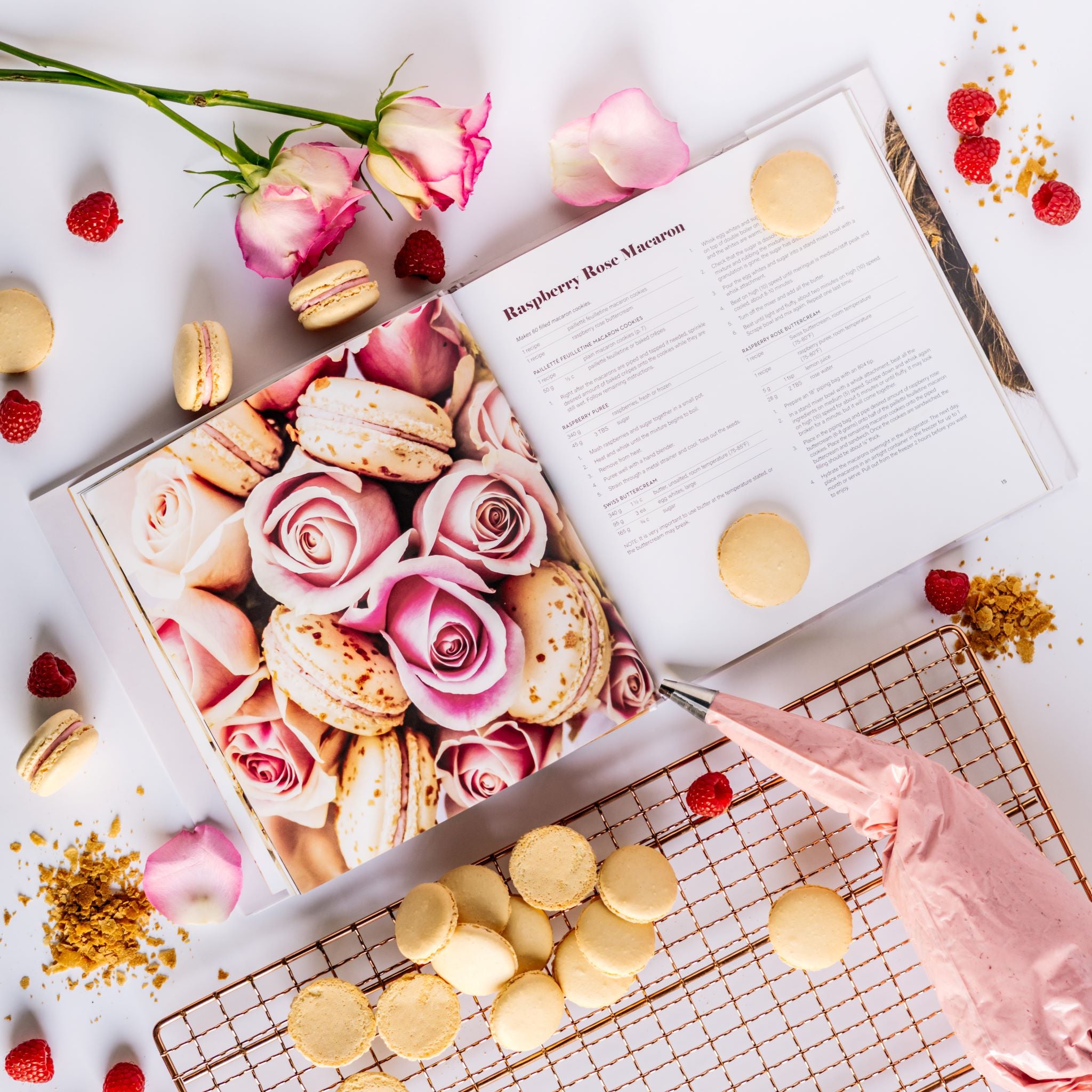 The Macaron Cookbook open to Raspberry Rose Macarons Page surrounded by roses, rose petals, macaron cookies, filled pastry bag and bakers cooling rack.